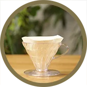 A circular image of a clear Hario V60 with a filter paper inside of it. The V60 is sitting on a wooden table.